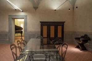 A dining area with a glass table and chairs, a piano in the background, and a decorative cabinet. - Stay in a prestigious place of interest in Florence in the city center (Firenze)