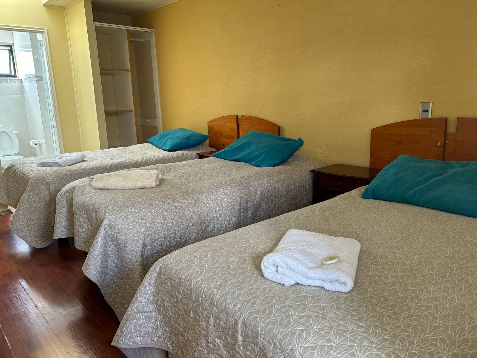 A hotel room with three beds, each with a grey patterned bedspread and a teal pillow. There is a wooden headboard on the right side of the room.