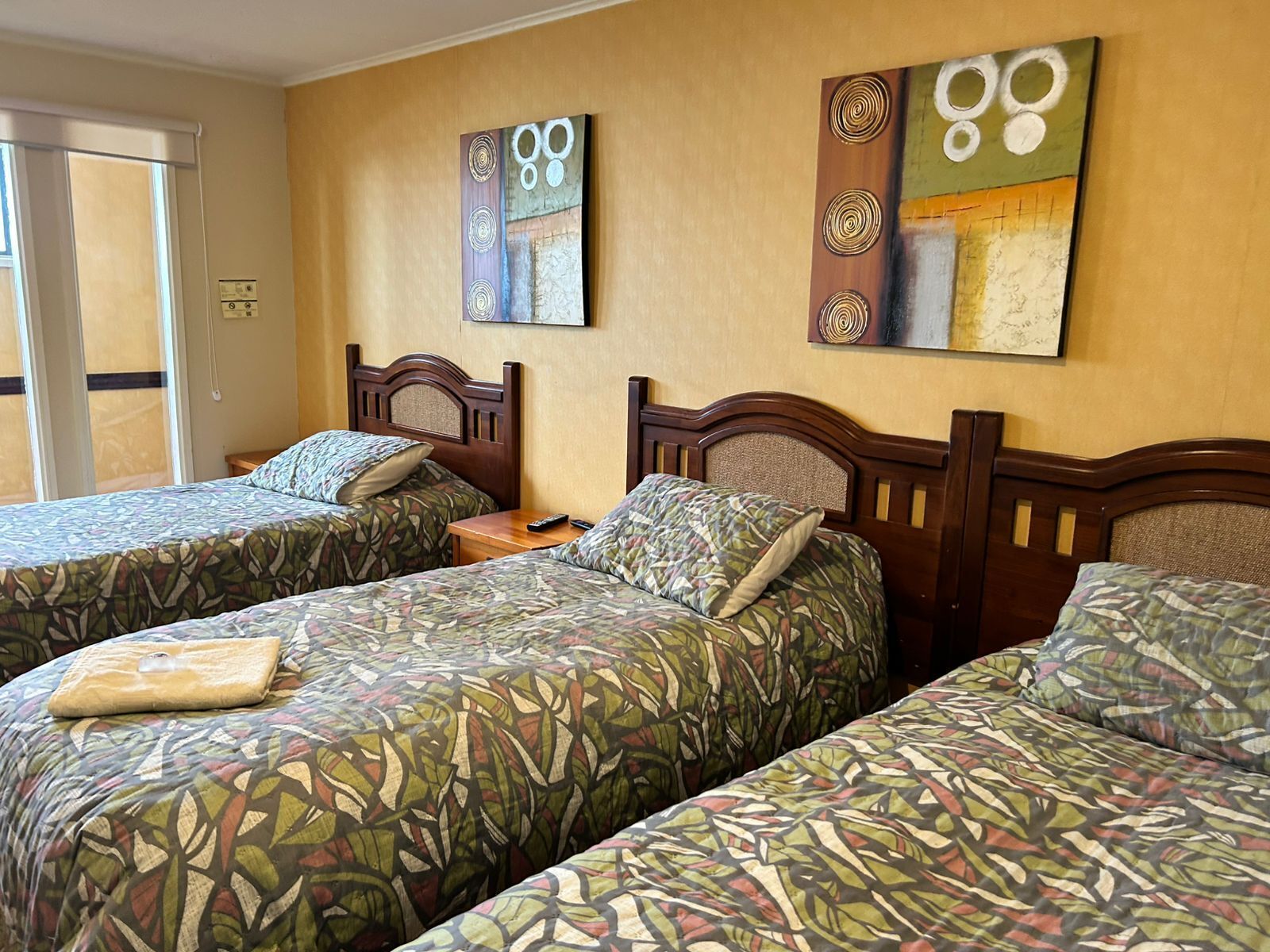 A hotel room with two single beds, a wooden headboard, a small bedside table, and a window with blinds.