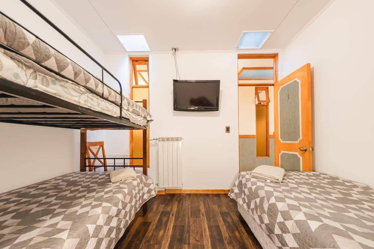 A dormitory room with bunk beds, a television, a radiator, and a door leading to another room.