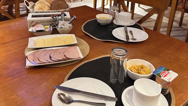 A breakfast setup with a variety of food items on a wooden table, including sliced ham, pastries, and cereal. - Hostal Don Guillermo (Natales)