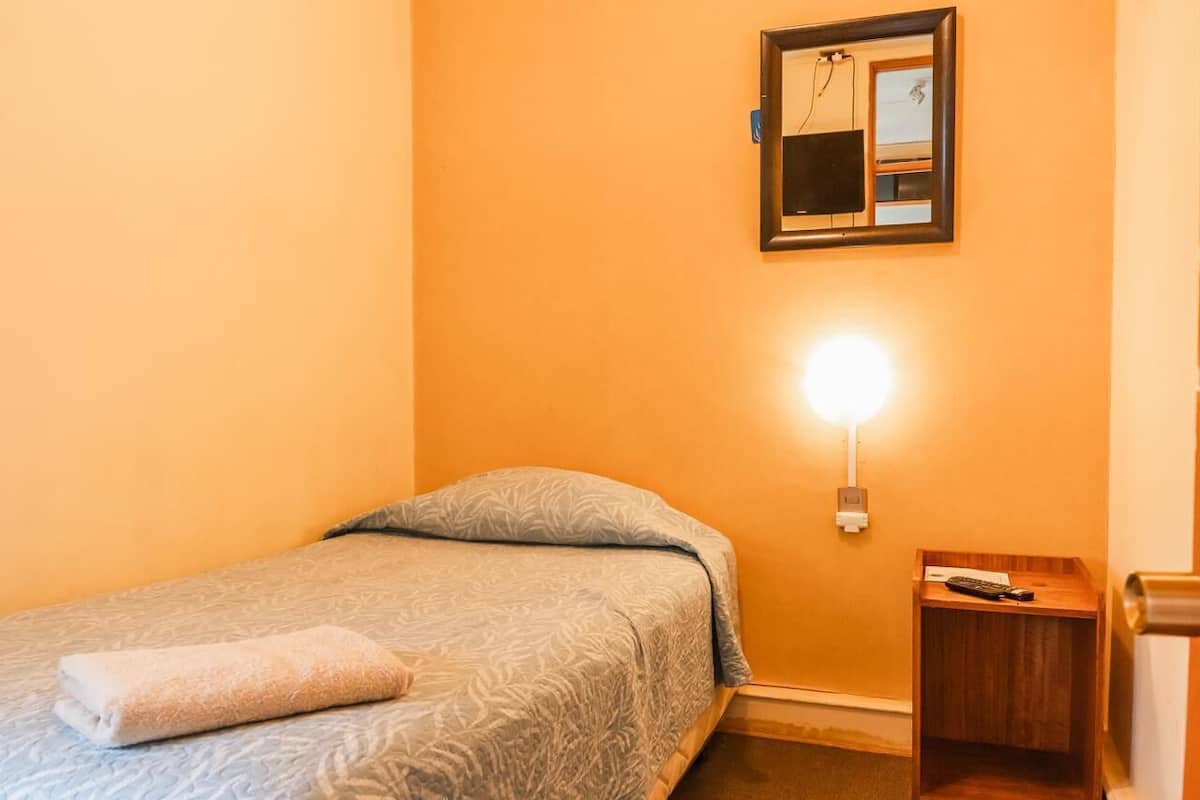 A single bed with a patterned bedspread, a bedside table with a remote control, and a wall-mounted light fixture.
