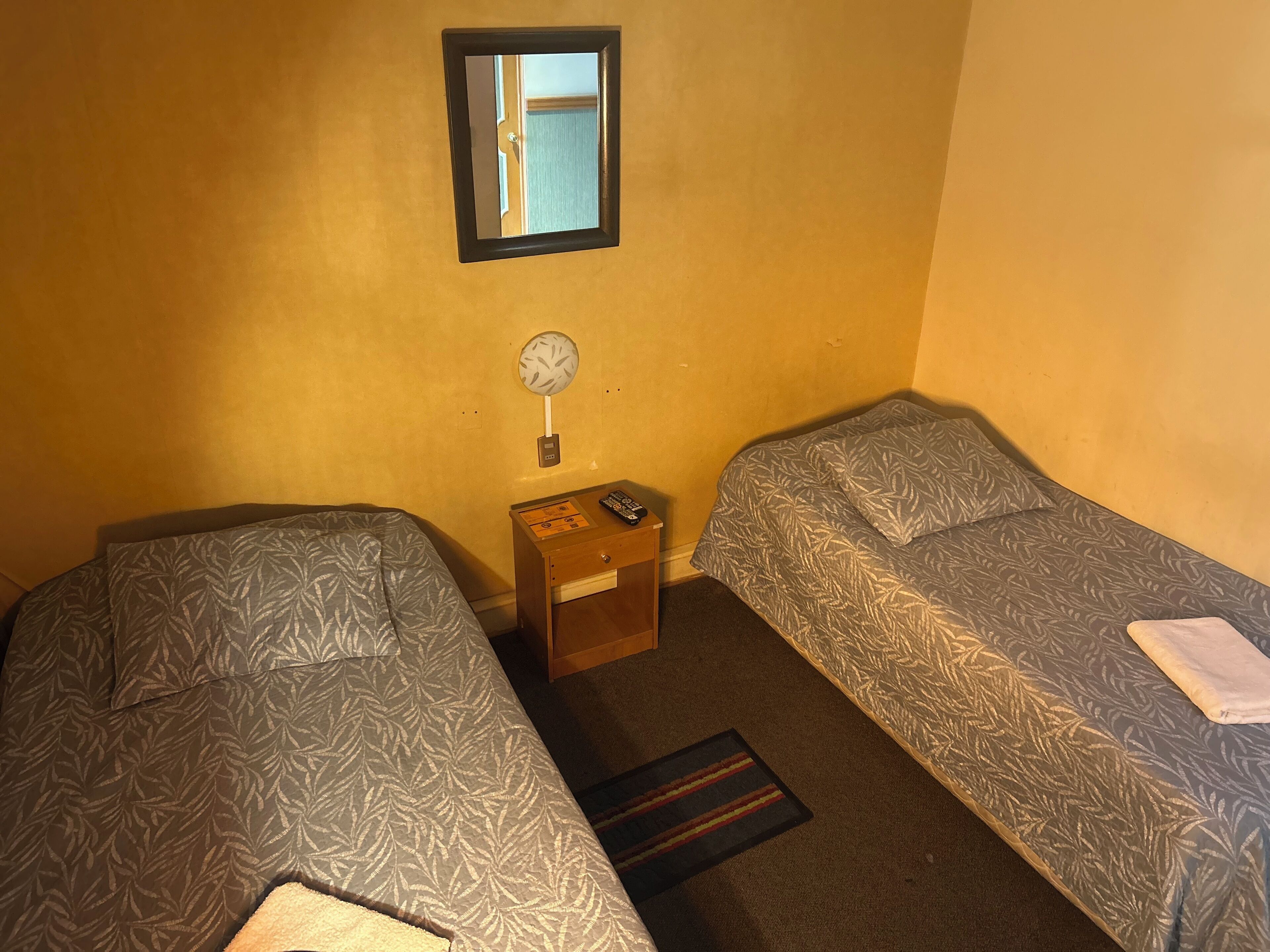 A small, single-room hotel room with two single beds, a bedside table with a lamp, a small wooden nightstand, and a mirror on the wall.