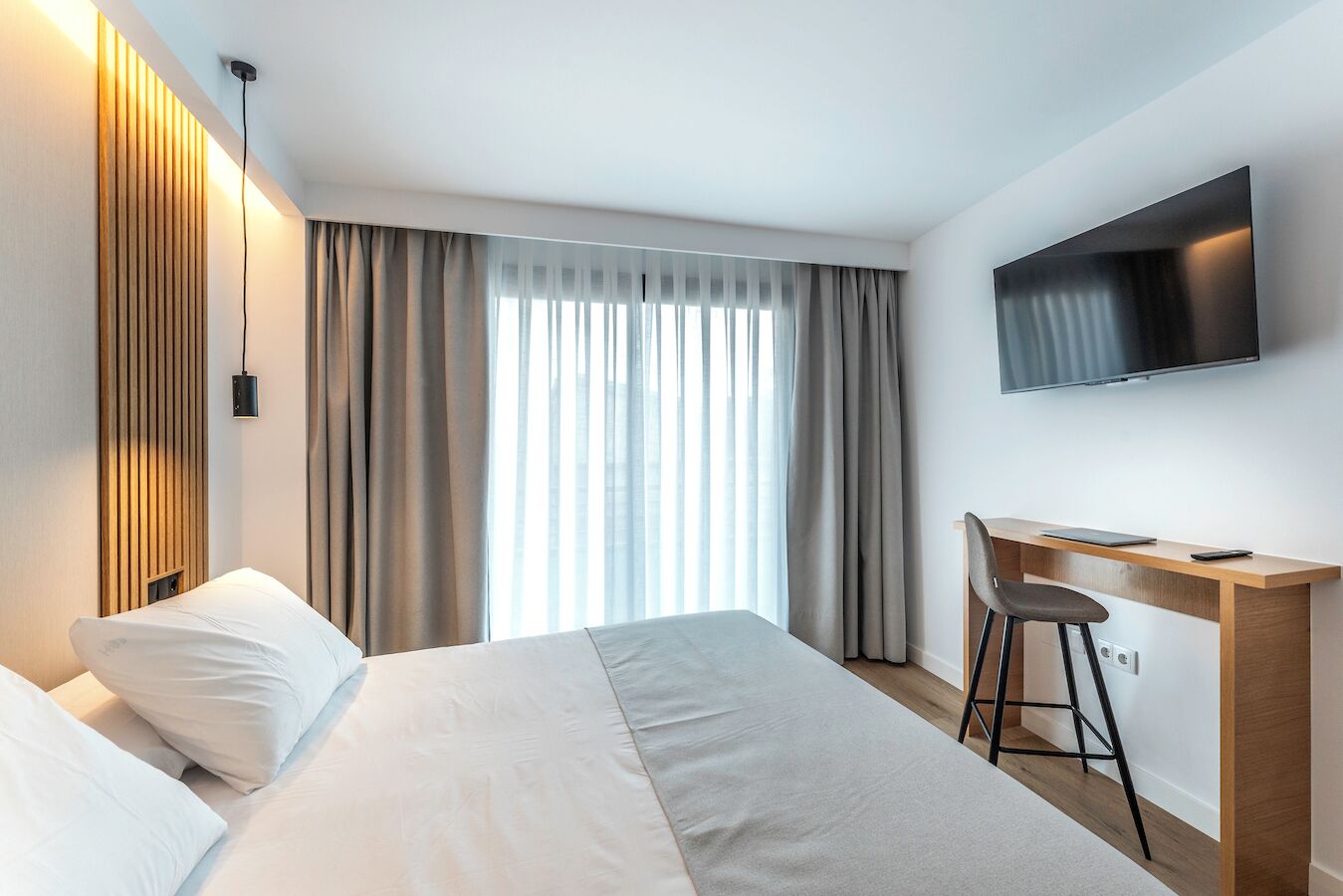 A hotel room with a bed, a desk, a chair, a TV, and a window with curtains.