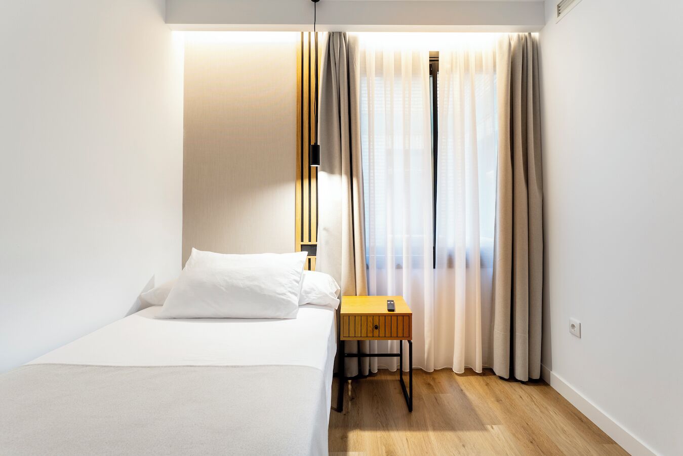 A hotel room with a bed, a bedside table with a remote control, and a window with sheer curtains.