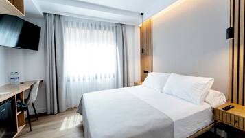 Habitación de hotel con cama, escritorio, televisión y ventana con cortinas.