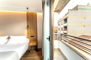 A hotel room with a bed, a wooden panel wall, a nightstand, and a balcony with a view of buildings.