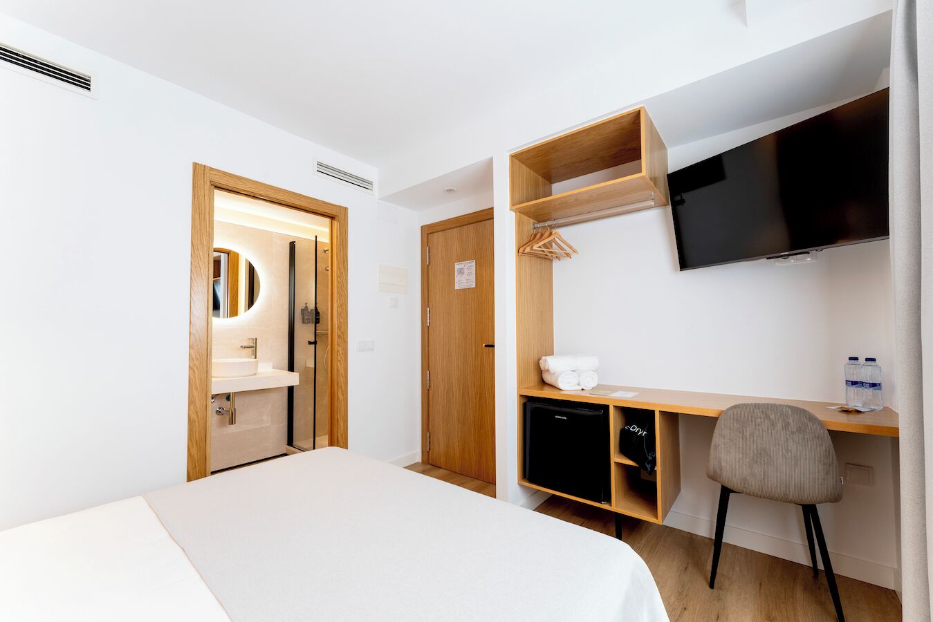 A hotel room with a bed, a desk with a chair, a TV, a mirror, and a wardrobe.