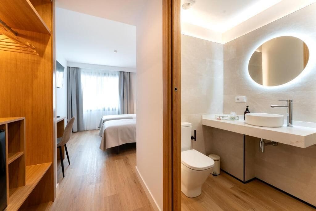 A modern hotel room with a wooden wardrobe, a bed, a bathroom with a round mirror, and a toilet.