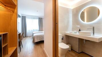 A modern hotel room with a wooden wardrobe, a bed, a bathroom with a round mirror, and a toilet.
