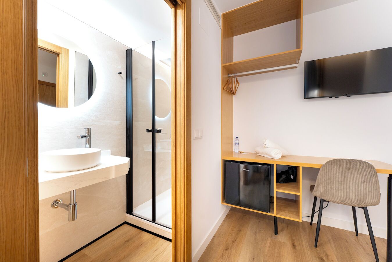 A modern bathroom with a sink, mirror, and glass shower enclosure. There is a wooden desk with a chair, a television mounted on the wall, and a shelf with a folded towel and a water bottle.