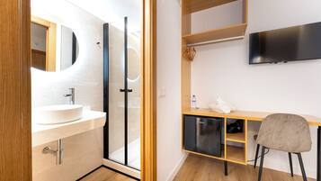 A modern bathroom with a sink, mirror, and glass shower enclosure. There is a wooden desk with a chair, a television mounted on the wall, and a shelf with a folded towel and a water bottle.
