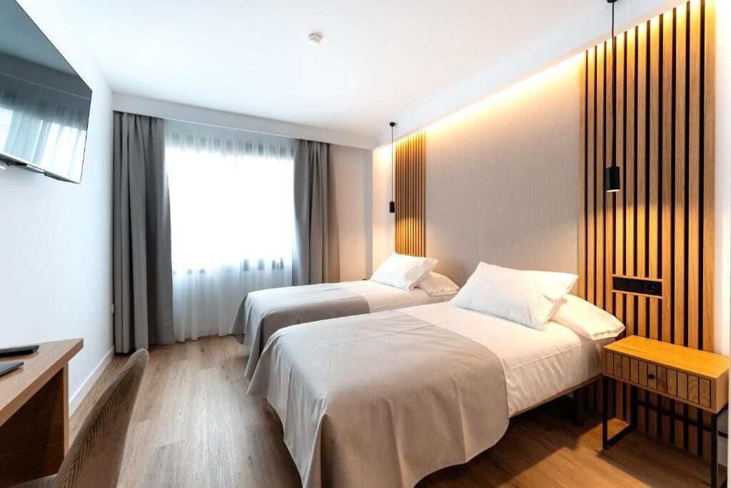 A hotel room with two beds, a wooden nightstand, a TV mounted on the wall, and a window with curtains.