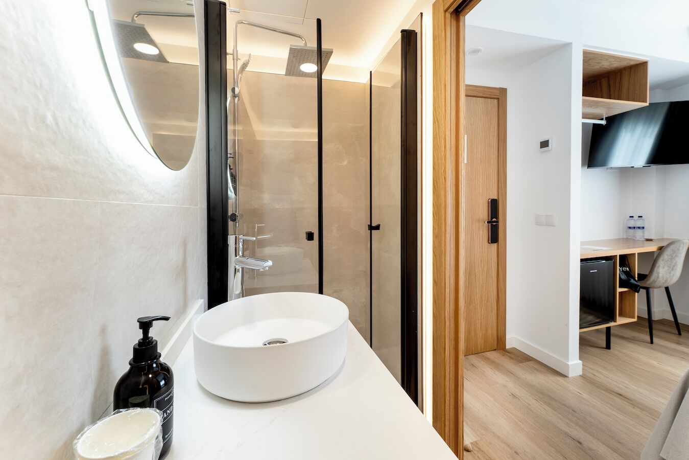 A modern bathroom with a large white sink, a glass shower enclosure, and a wooden door leading to a bedroom with a TV and a desk.