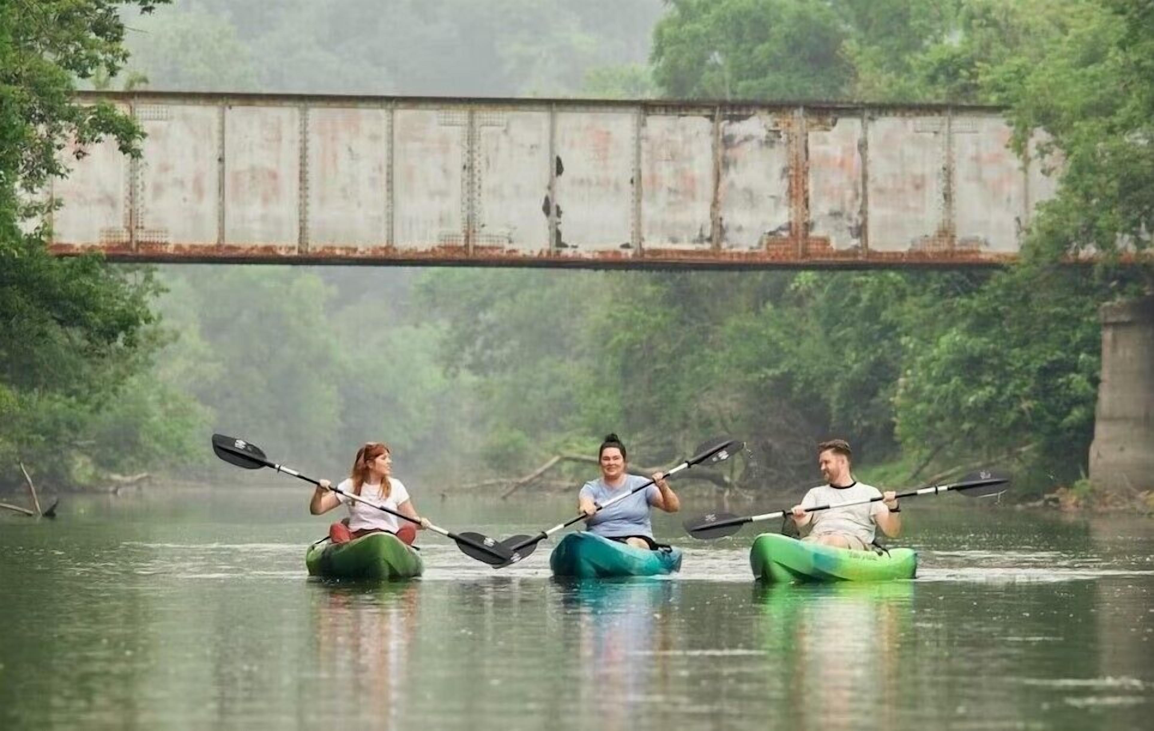 Tres kayakistas en un río tranquilo bajo un puente.