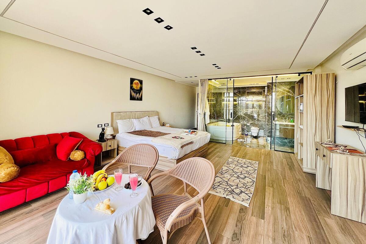 A modern hotel room with a red sofa, a bed, a dining area with a table and chairs, and a bathroom with a bathtub and shower.