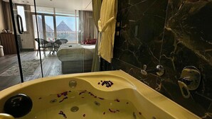 A luxurious bathroom with a marble wall, a bathtub with rose petals, and a view of a cityscape through large windows. - Roone Pyramids View Inn (Giza)