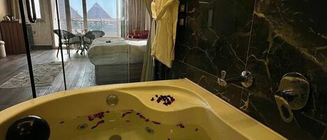 Un baño lujoso con pared de mármol, una bañera con pétalos de rosa y vistas a la ciudad a través de amplias ventanas.