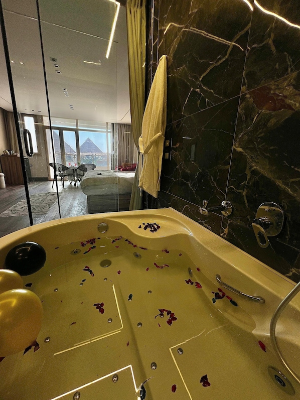 A luxurious bathroom with a marble wall, a bathtub with rose petals, and a view of a cityscape through large windows. - Roone Pyramids View Inn (Giza)