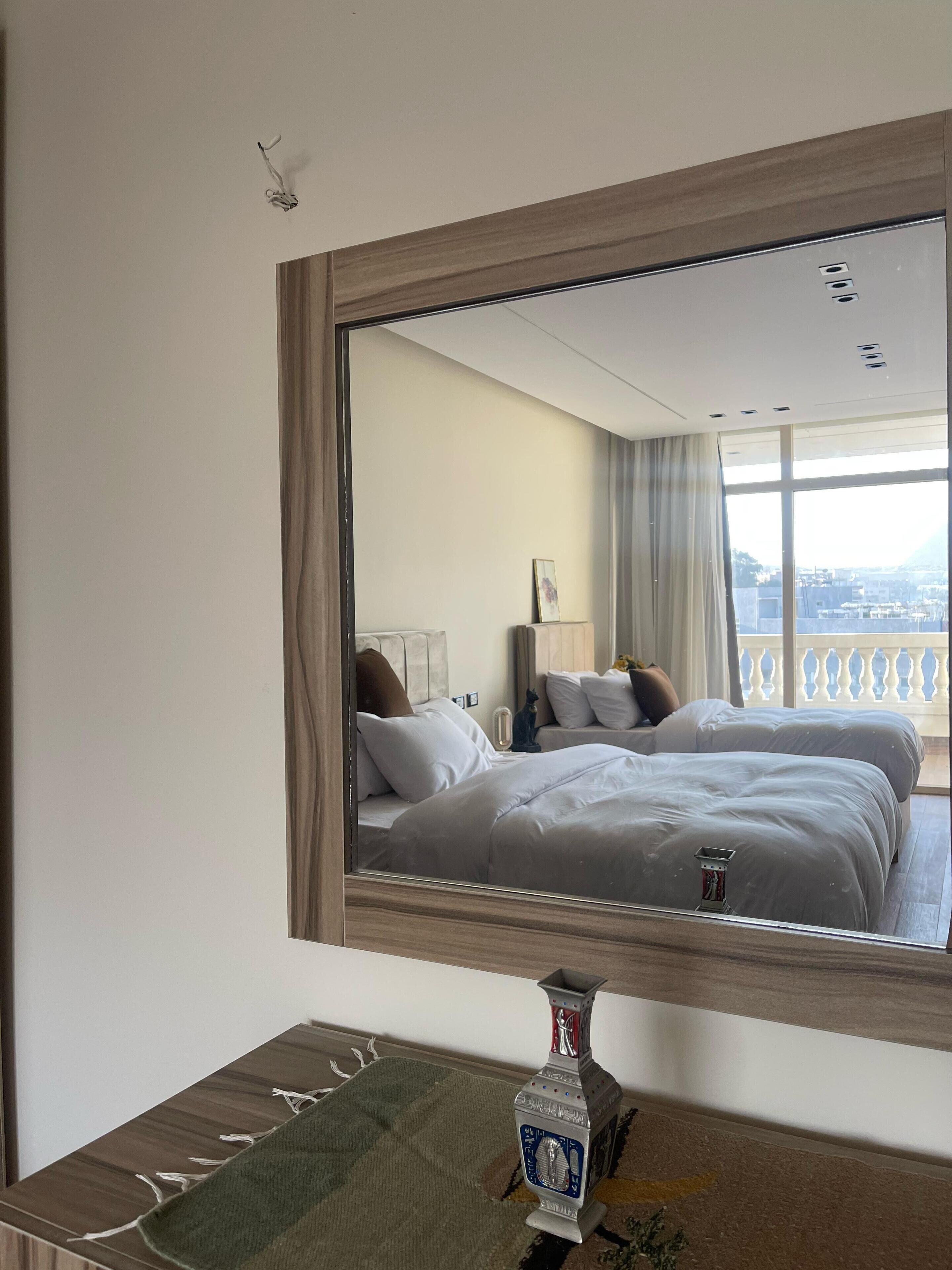 Een hotelkamer met een grote spiegel die een bed, een nachtkastje en uitzicht op de stad door grote ramen weerspiegelt.