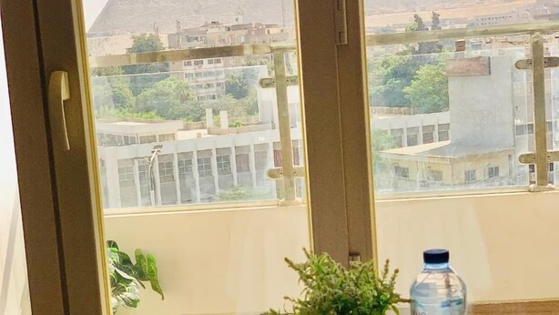 A table with drinks, fruits, and a water bottle, with a view of pyramids through a window. - Roone Pyramids View Inn (Giza)