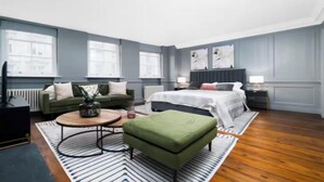 Una habitación de hotel moderna con un sofá verde, una mesa de centro redonda, un reposapiés verde, una cama con edredón blanco y un ventanal grande.