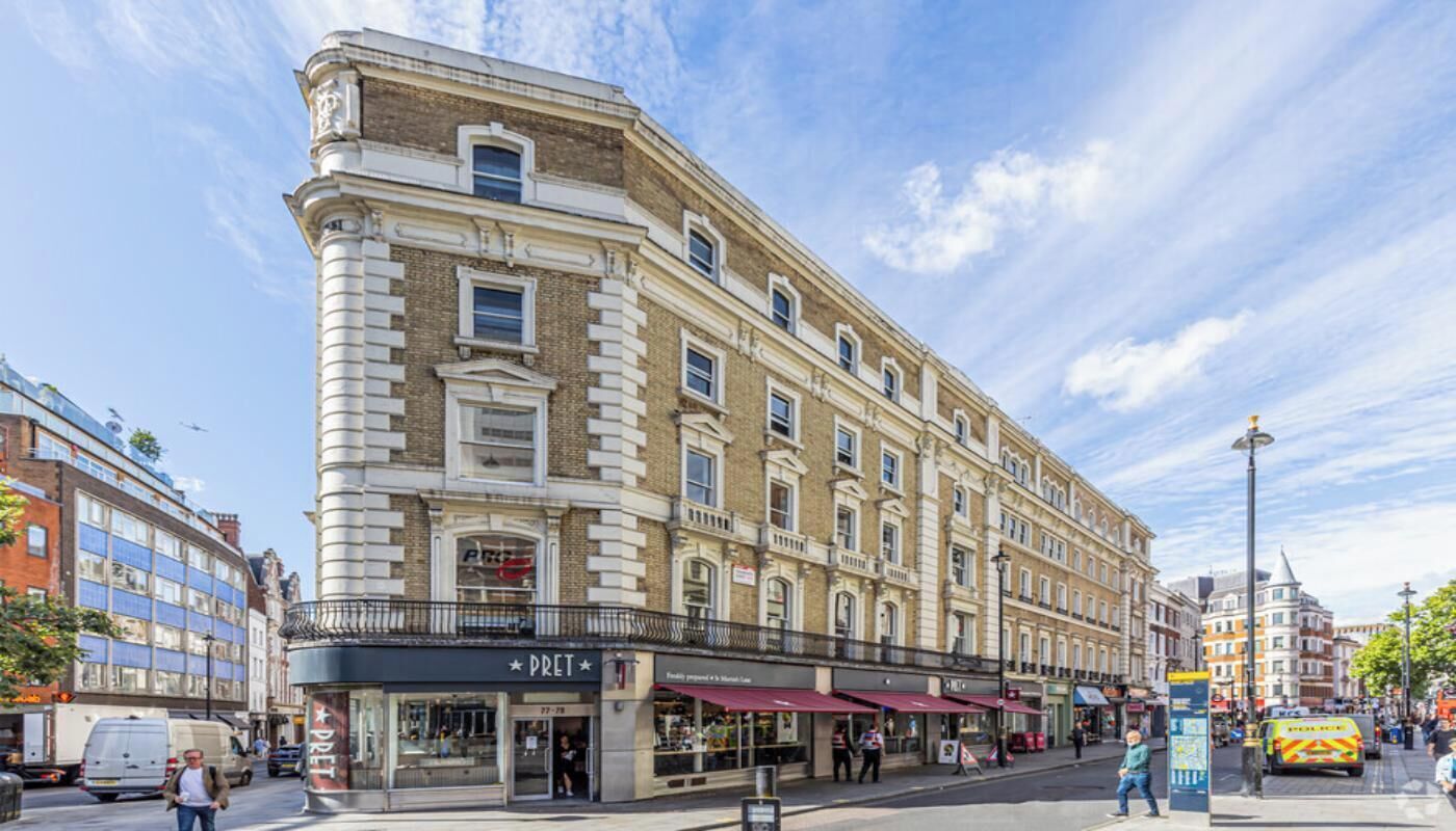 Um edifício histórico com entrada de canto, fachada visível com uma placa e rua com pedestres e veículos.