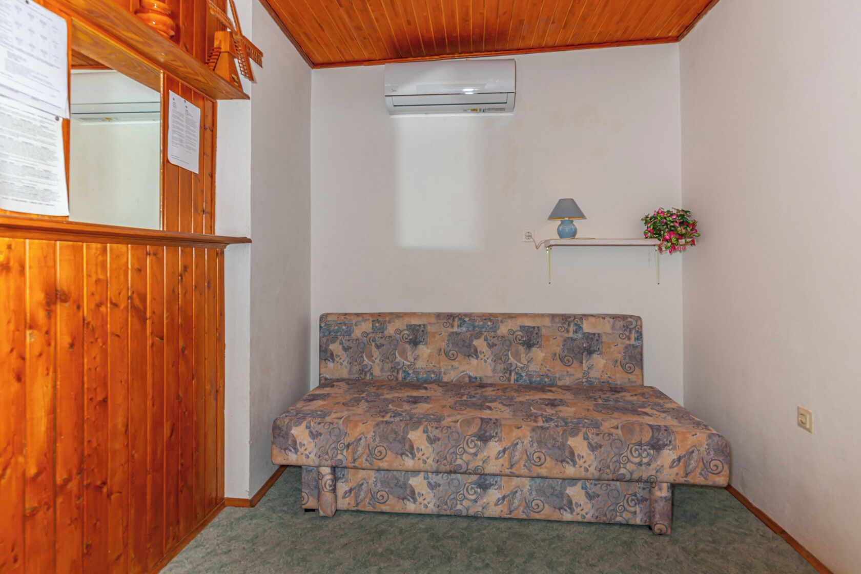 A small room with a patterned sofa, a wooden cabinet, an air conditioning unit, and a small shelf with a lamp and flowers.