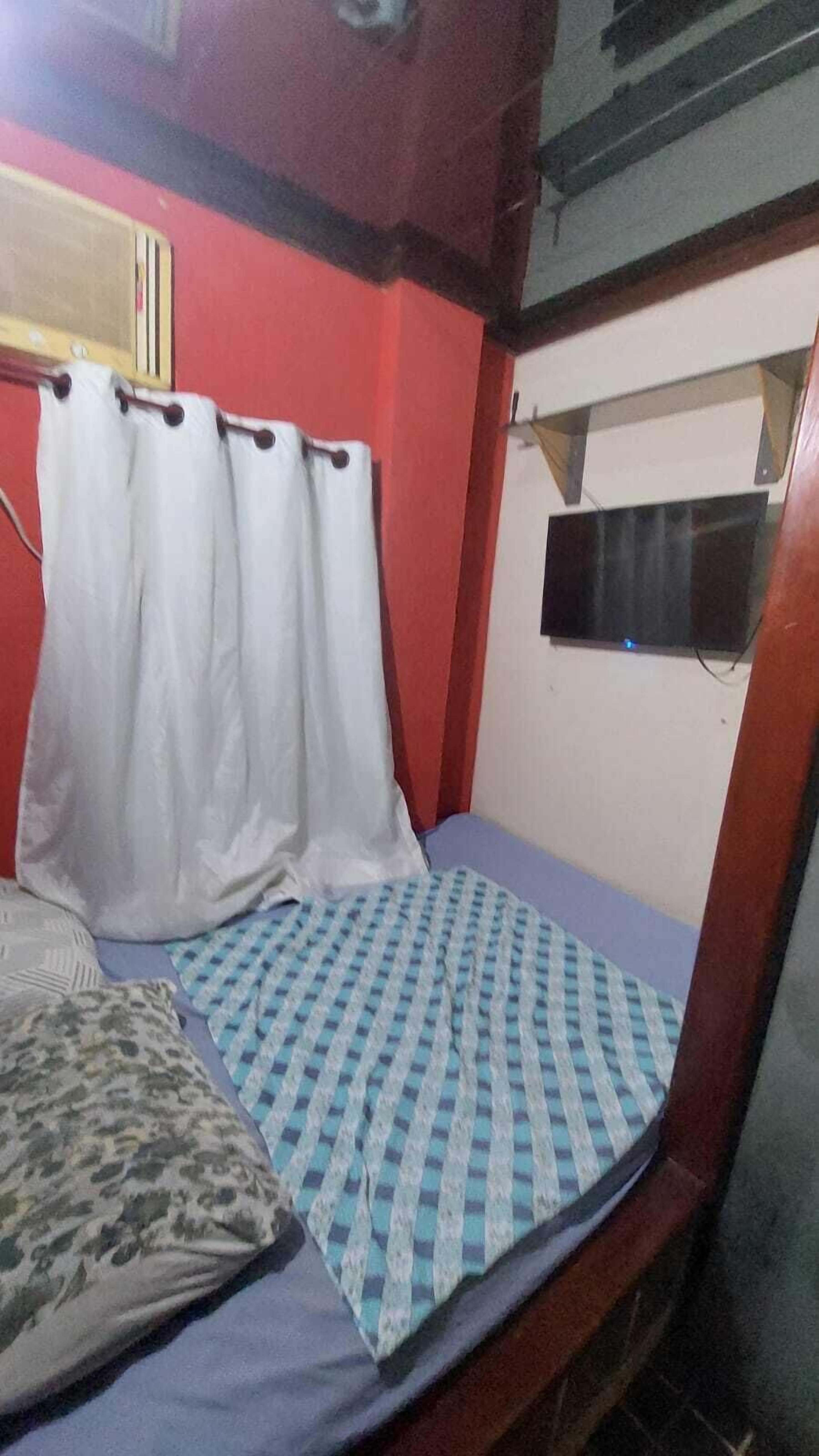 Ein kleines Zimmer mit einem Einzelbett, einer karierten Bettdecke, einem weißen Vorhang und einem kleinen Fenster.