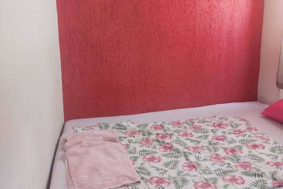 A small, single-bed room with a red textured wall, a floral bedspread, and a pink towel on the bed.
