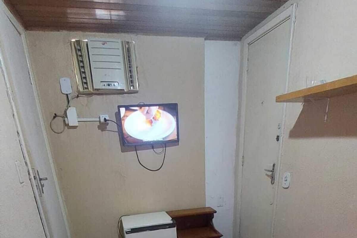 A small, compact room with a bed, a small desk, a television mounted on the wall, and a heater.