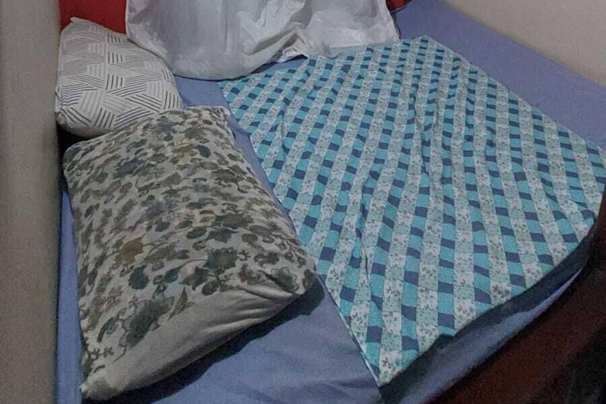 A small, single-bed room with a red wall, a white curtain, a patterned bedspread, and a small air conditioning unit mounted above the bed.