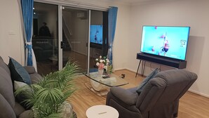 Un salon comprenant un canapé, une table basse ornée d’un vase de fleurs, un téléviseur diffusant un événement sportif et une porte-fenêtre coulissante donnant sur une autre pièce.