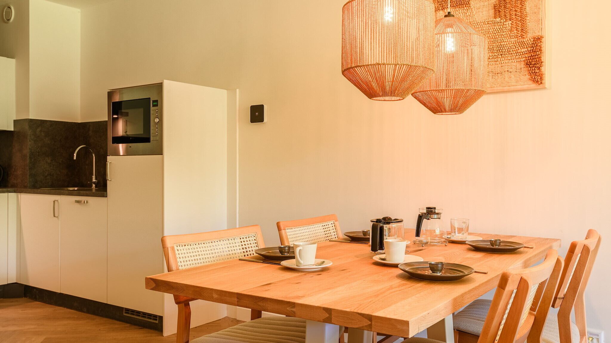 Una cocina moderna con microondas integrado, un fregadero y un refrigerador. Hay una mesa de comedor de madera con sillas y una cafetera encima.