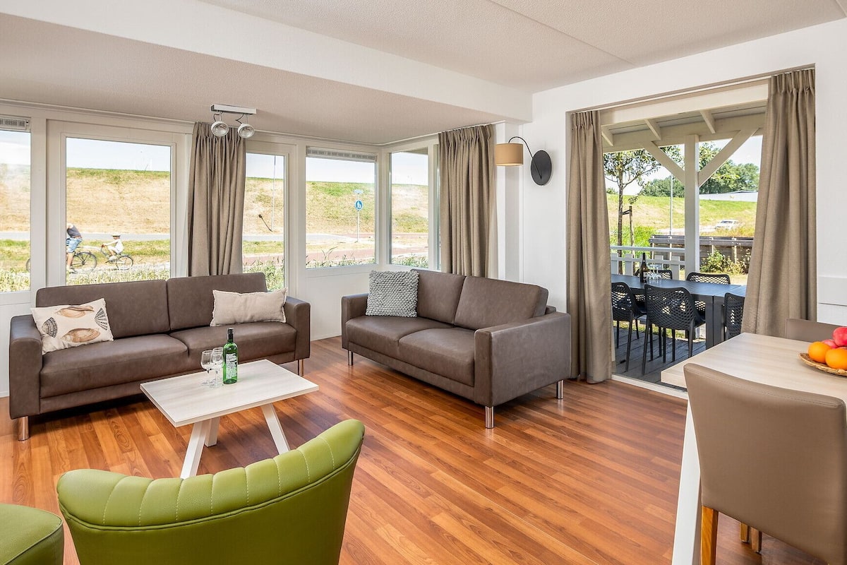 A modern living room with two sofas, a coffee table, and a dining area with a table and chairs. Large windows offer views of the outdoors.