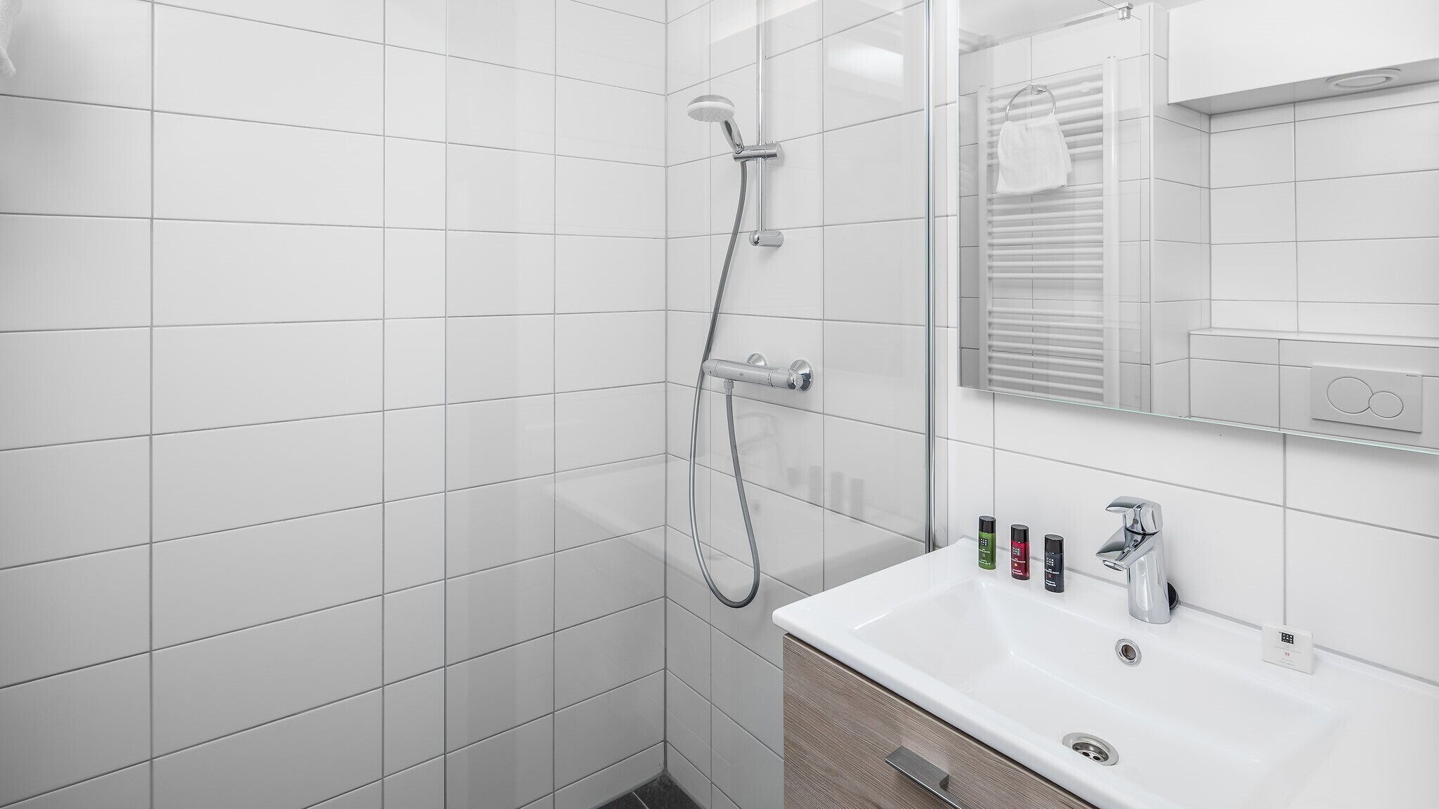 A modern bathroom with white tiles, a glass shower enclosure, a white sink with a mirror, and toiletries on the countertop.