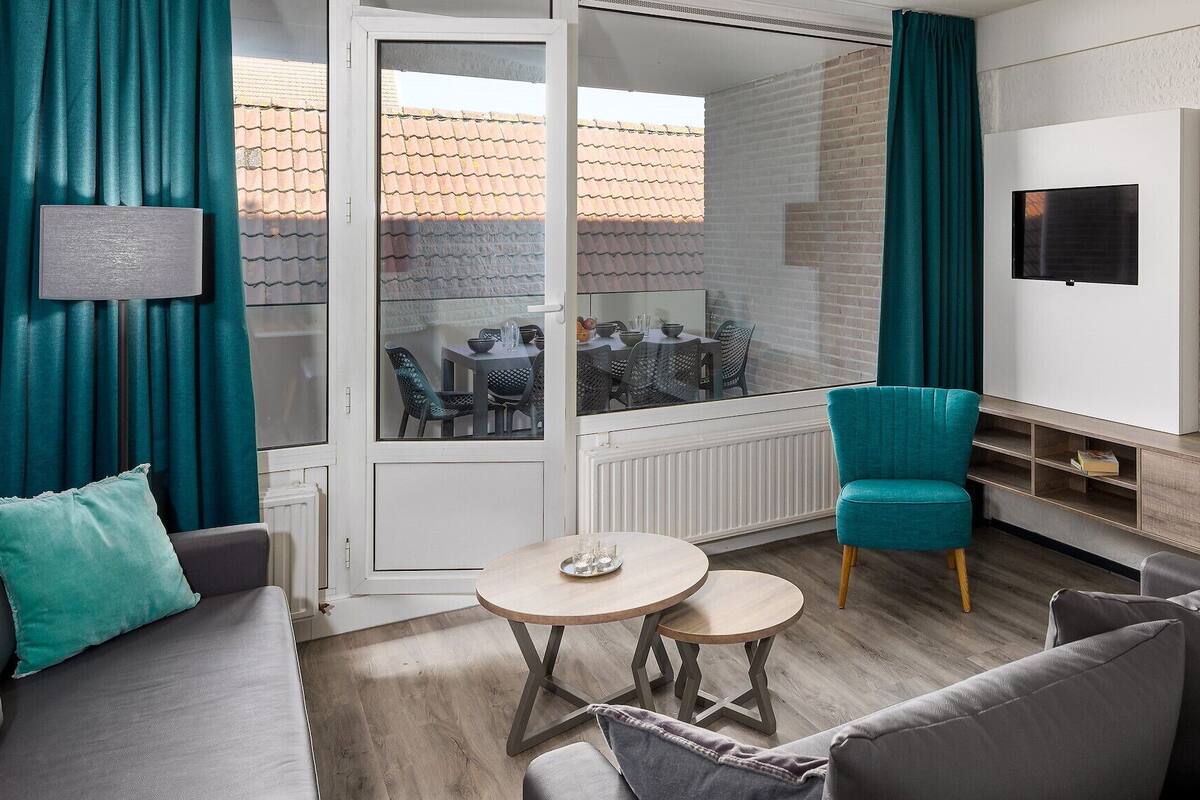 A modern living room with a sofa, two small tables, a teal armchair, and a TV mounted on the wall.