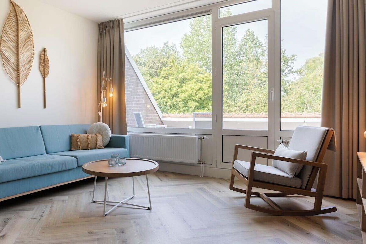 A modern living room with a blue sofa, a wooden rocking chair, a coffee table, and large windows offering a view of trees.