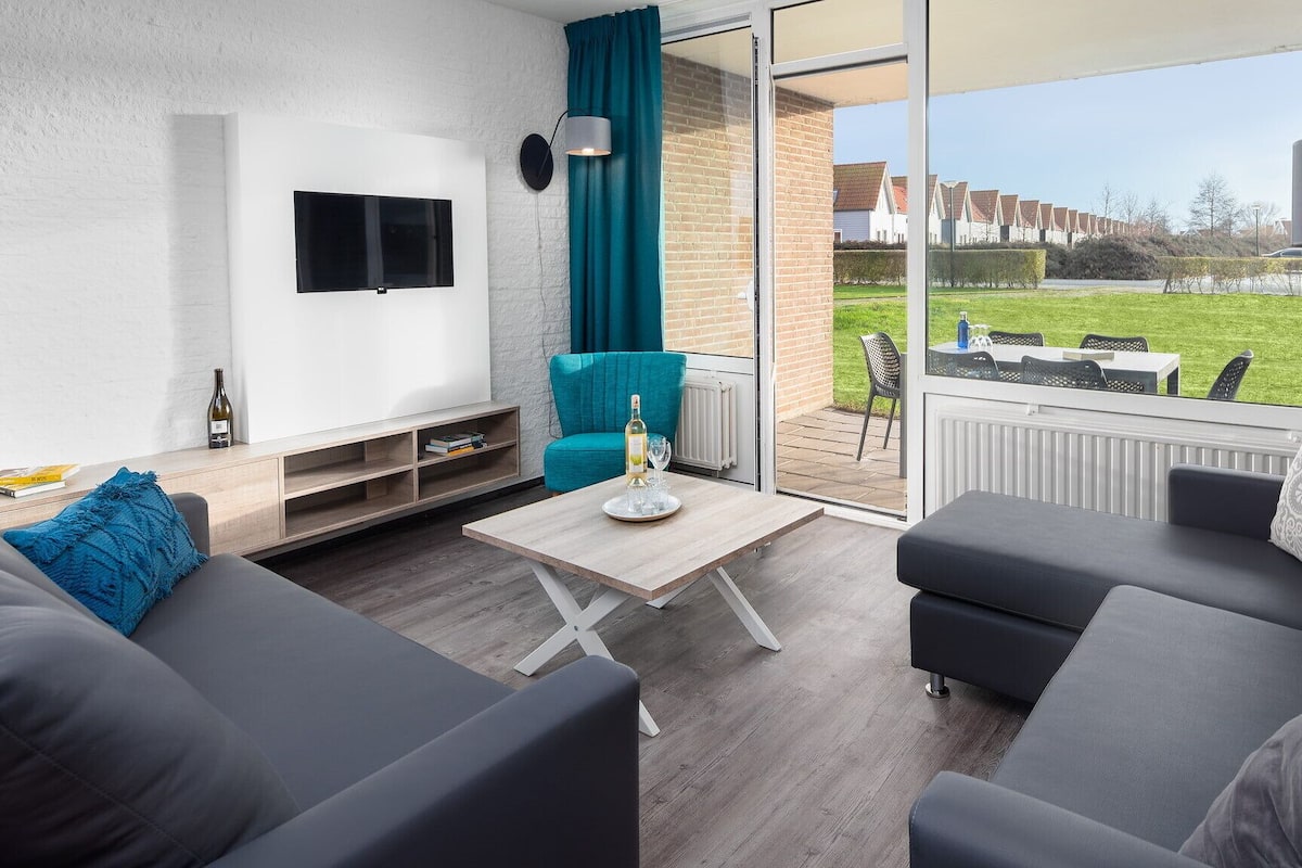 A modern living room with a sofa, a coffee table, and a TV. There is a sliding glass door leading to an outdoor patio with a table and chairs.