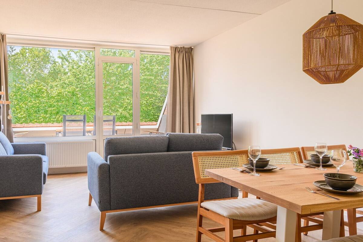 A modern living room with a sofa set, a dining table with chairs, and a large window offering a view of greenery outside.