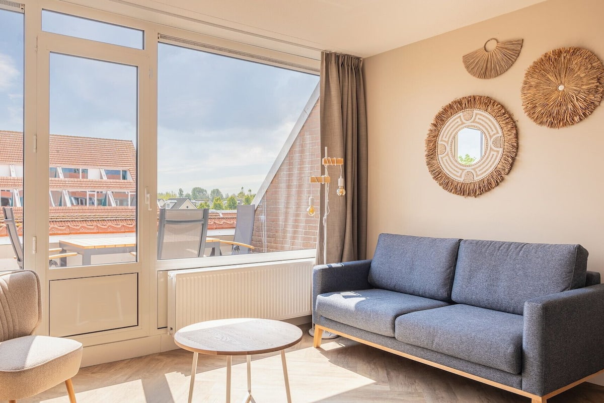 A modern living room with a grey sofa, a round table, and two chairs. There is a large window with a view of buildings and a clear sky.