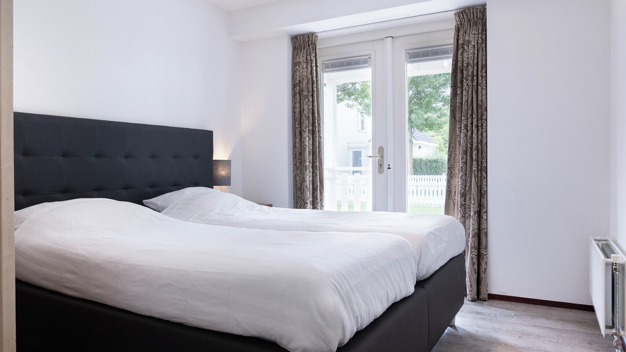 A bedroom with a large bed, a dark headboard, and a view of a white house through the window.