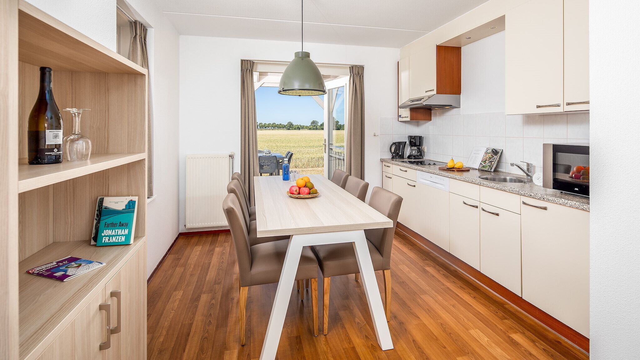 Een moderne keuken met witte kasten, een eettafel met stoelen, een hanglamp en uitzicht op een veld door het raam.