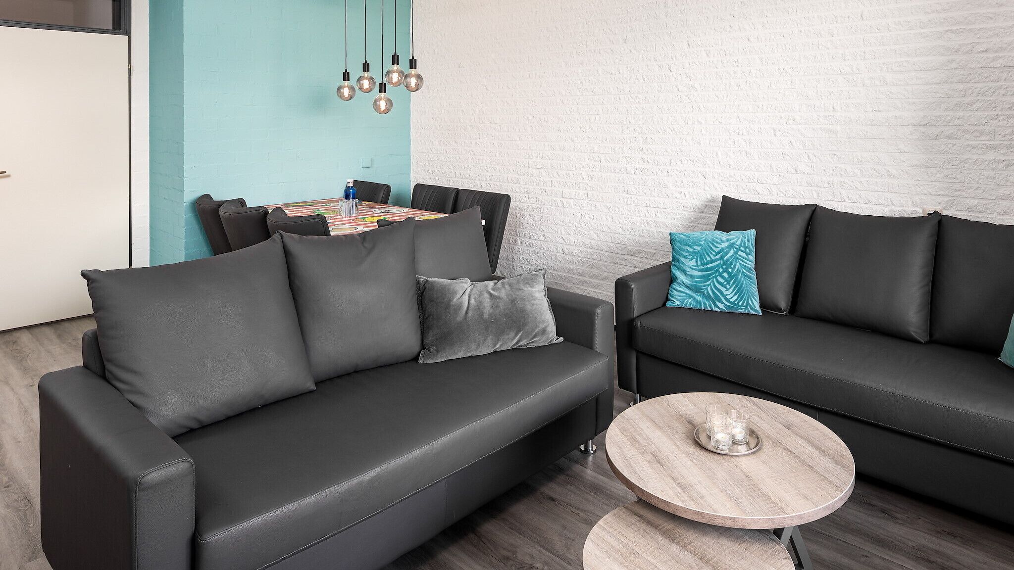 A modern living room with two black sofas, a round wooden coffee table, and a dining area with a colorful tablecloth.