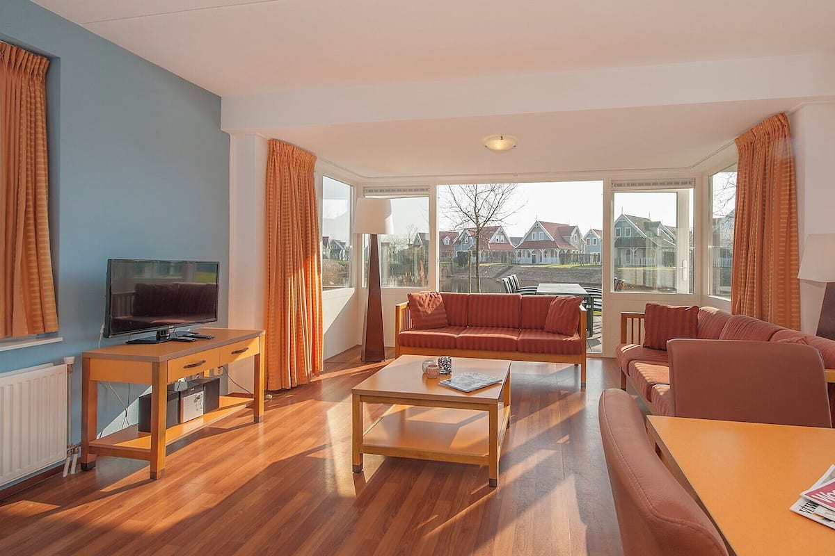 A modern living room with a TV, sofa, coffee table, and a view of houses through large windows.
