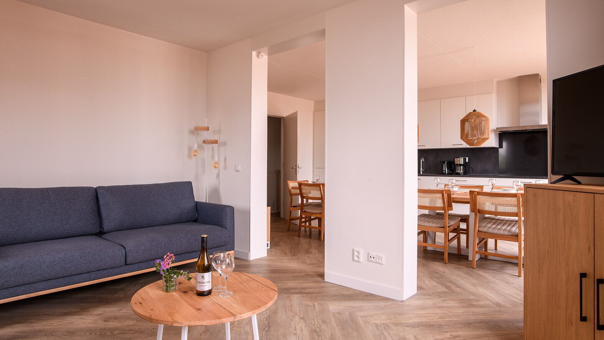 A modern living room with a sofa, a dining area with chairs, a kitchen with a TV, and a wooden coffee table with a bottle and glasses.