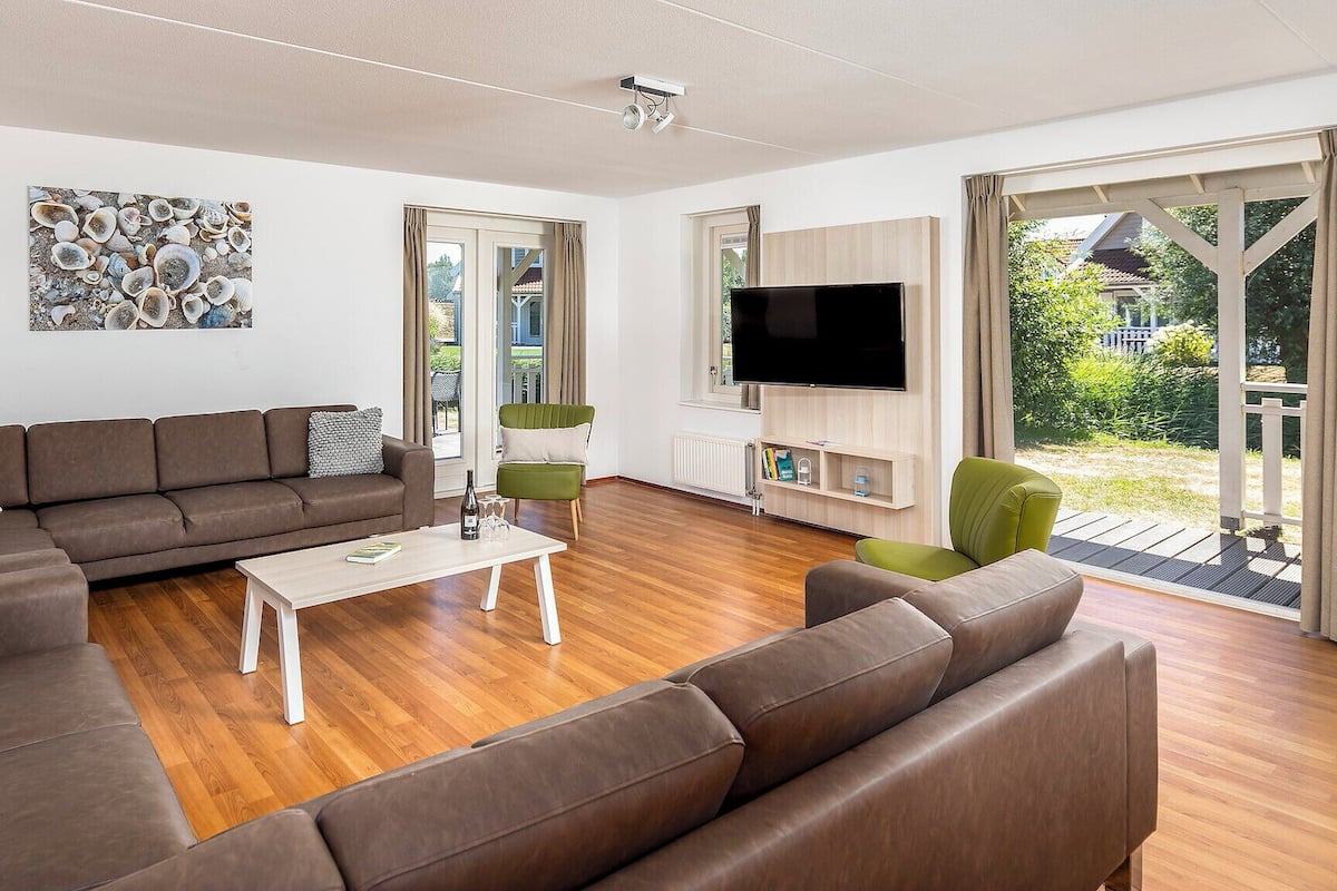 A modern living room with a brown leather sofa, a white coffee table, and a green armchair. There is a flat-screen TV mounted on the wall and a painting on the wall.