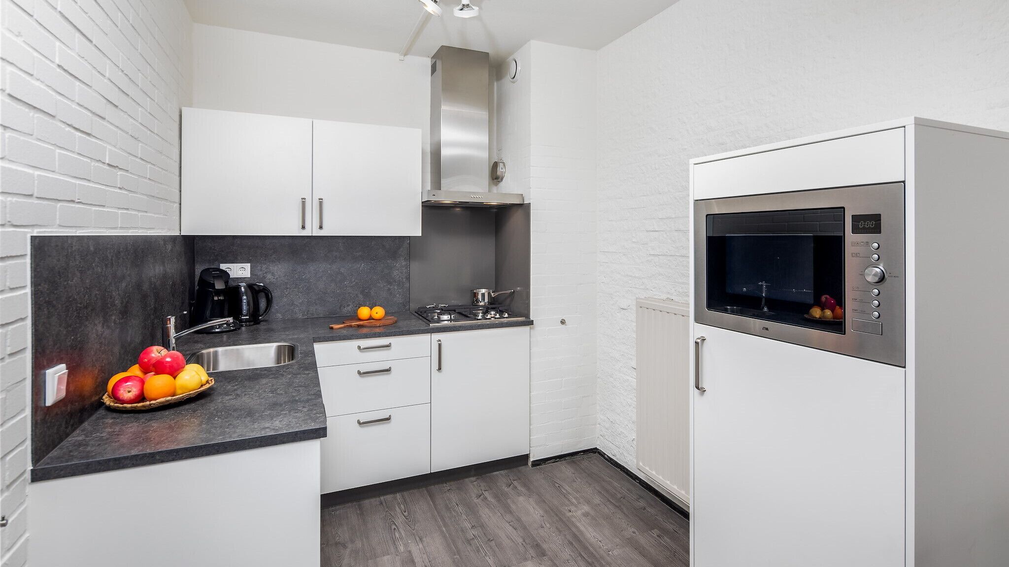 Una cucina compatta con mobili bianchi, forno integrato, macchina del caffè e ciotola di frutta.