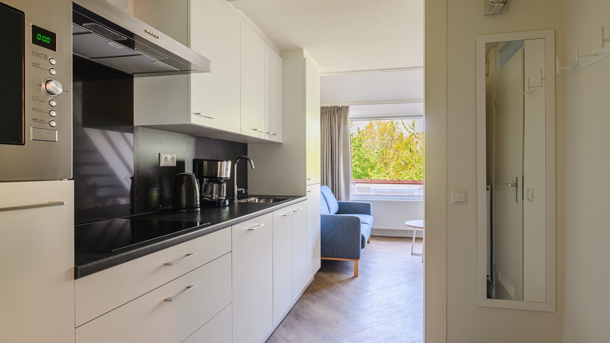 Una cocina moderna con armarios blancos, una encimera negra y una cafetera. Se ve una ventana con cortinas, una silla azul y una puerta que da a otra habitación.