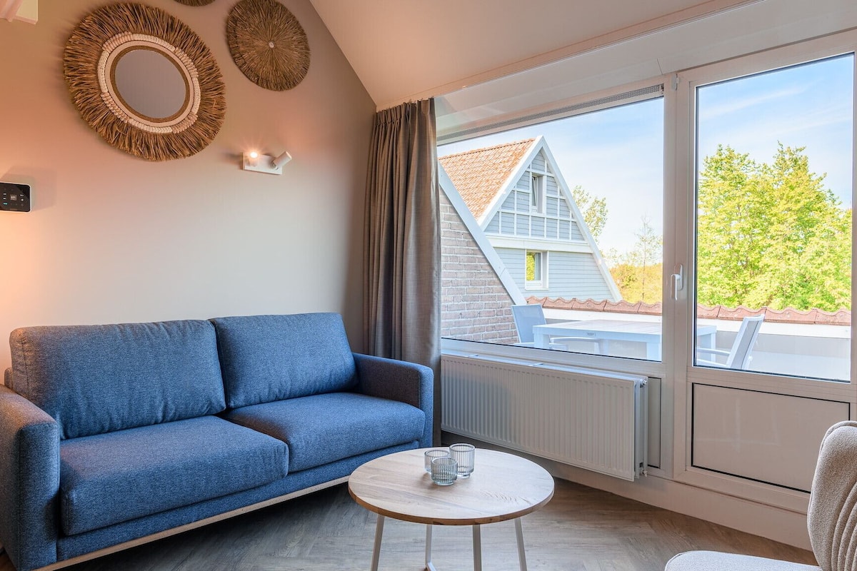 A modern living room with a blue sofa, a round wooden coffee table, and a large window offering a view of a house and trees.
